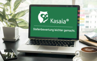 Laptop auf Schreibtisch mit grünem Kasaia®-Screen „Stellenbewertung leichter gemacht“, daneben Pflanze, Smartphone und Kaffeetasse.